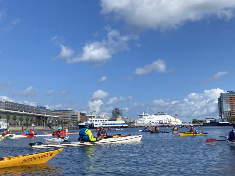 Teilnehmende der Schwentinewanderfahrt in ihren Booten auf der Kieler Förde, im Hintergrund die Schwedenfähre.